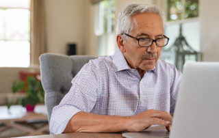Senior man working with laptop at home. Old man using computer at home sitting on chair and looking at screen. Elderly grandfather wearing eyeglasses and working on laptop in living room.